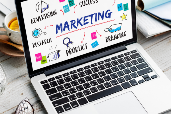 A top-down view of an open laptop on a white wooden desk. The screen displays a creative "Marketing" mind map with hand-drawn illustrations and colorful icons for advertising, research, branding, and product development. A cup of tea and a pair of glasses sit beside the laptop.